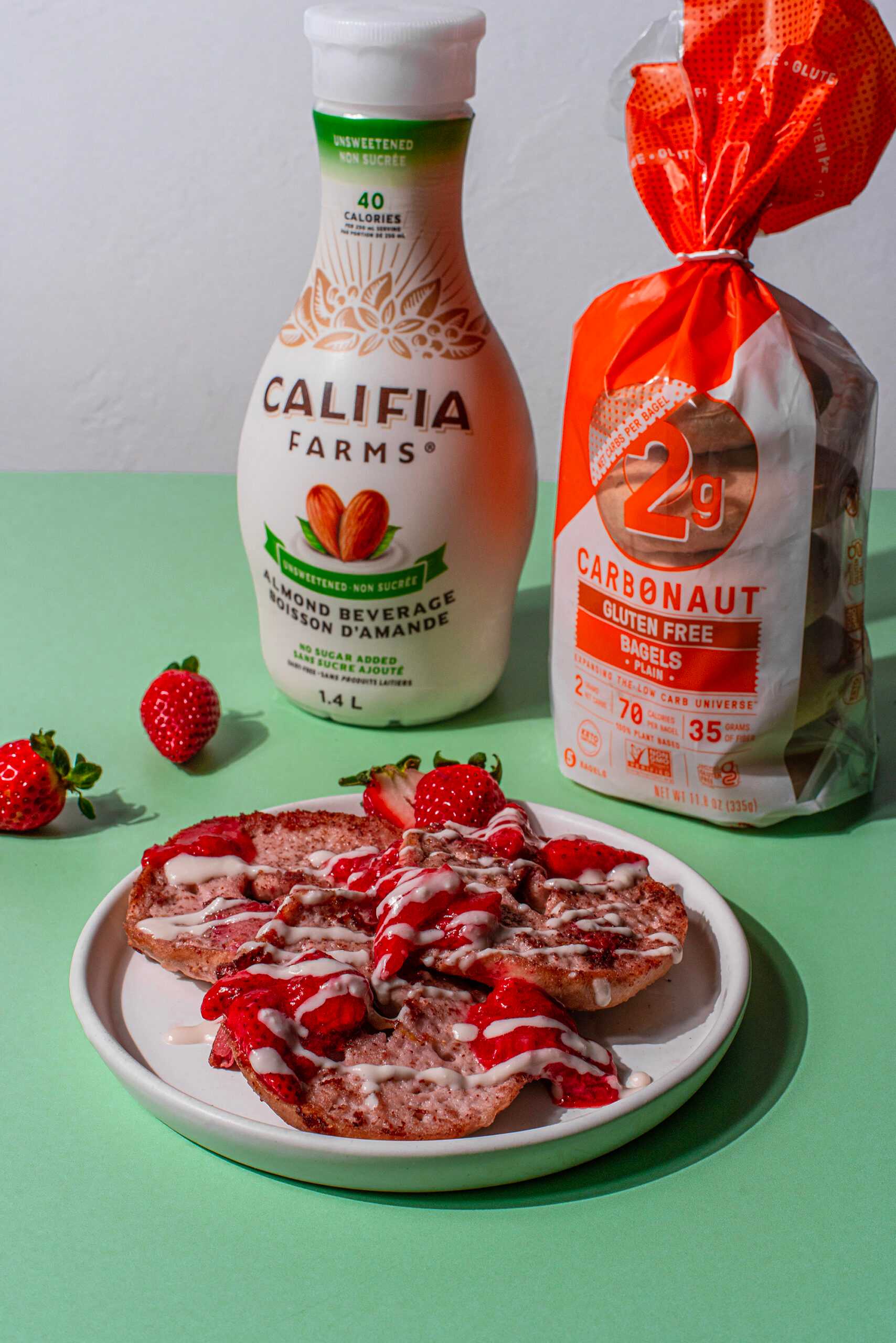 Strawberry bagel french toast sits in the center of the image, with Califia Farms Unsweetened Almond Beverage and Carbonaut Gluten-Free, Low-Carb Plain Bagels in the background.