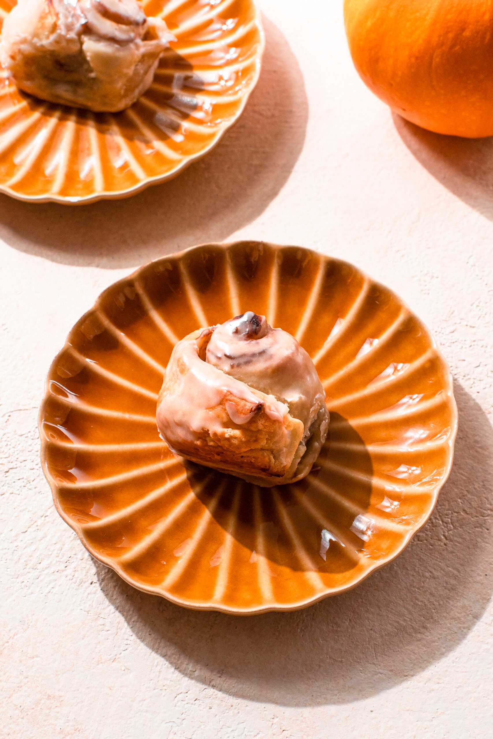 A pumpkin-spice flavored cinnamon roll sits in the center of the image on a decorative plate.